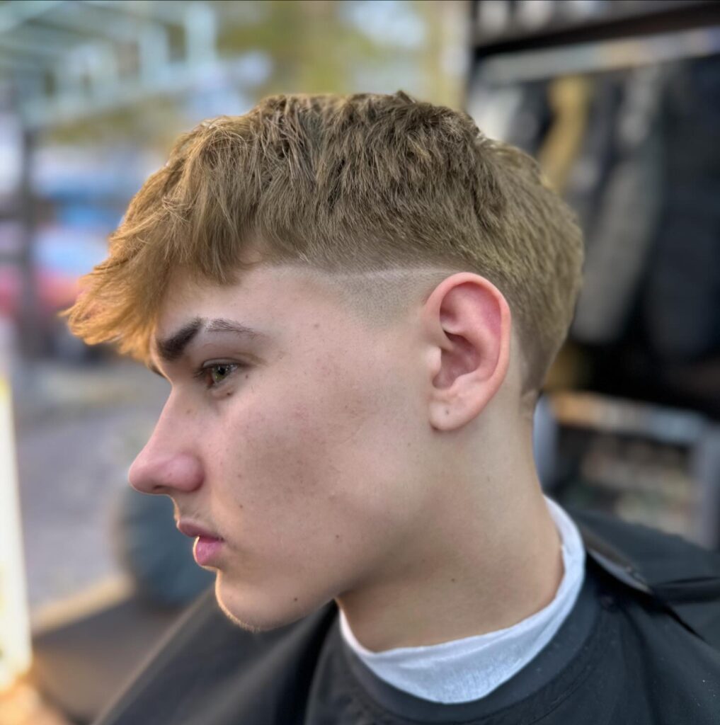 Side profile of a young man with a mid taper fade haircut and textured fringe, showing short faded sides and longer, tousled hair on top.