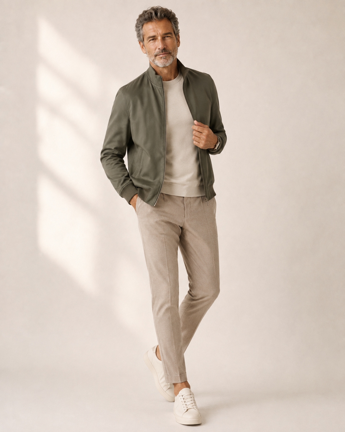 Modern casual outfit for a man over 50 with a matte olive bomber, stone knit, taupe trousers and white sneakers